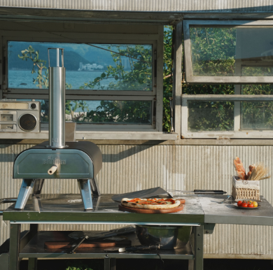 A portable pizza oven set up on a cart in front of a vintage camper.