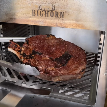 A cooked tomahawk steak being removed from a BIGHORN salamander broiler with metal tongs.