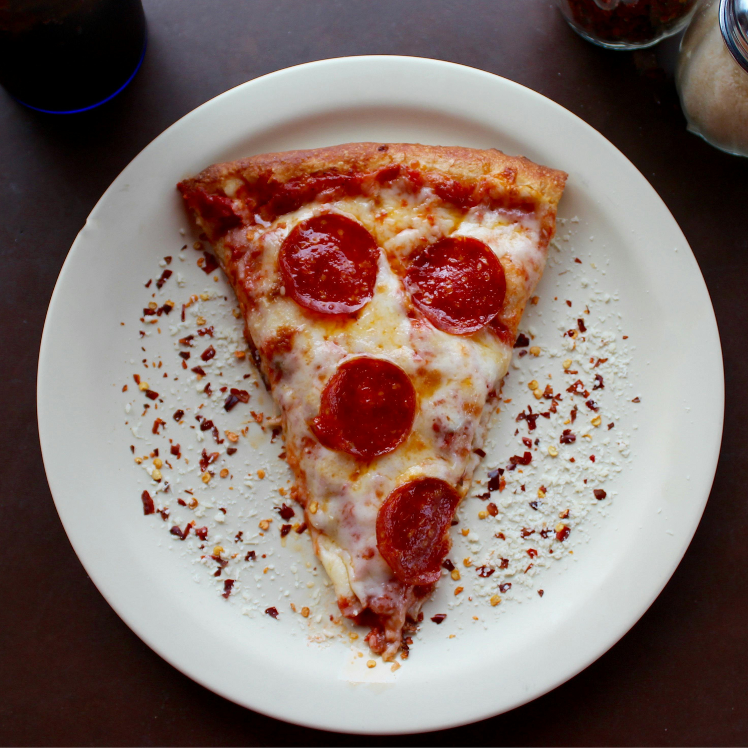 A slice of pepperoni pizza on a white plate, sprinkled with red pepper flakes and grated cheese.
