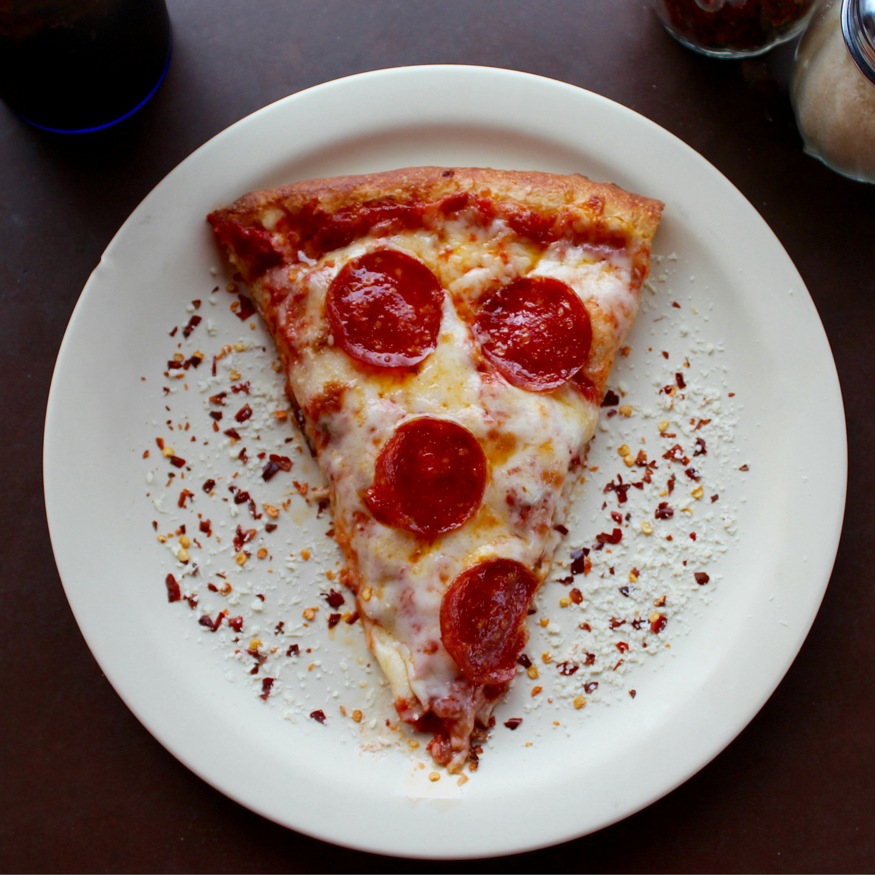 A slice of pepperoni pizza on a white plate, sprinkled with red pepper flakes and grated cheese.