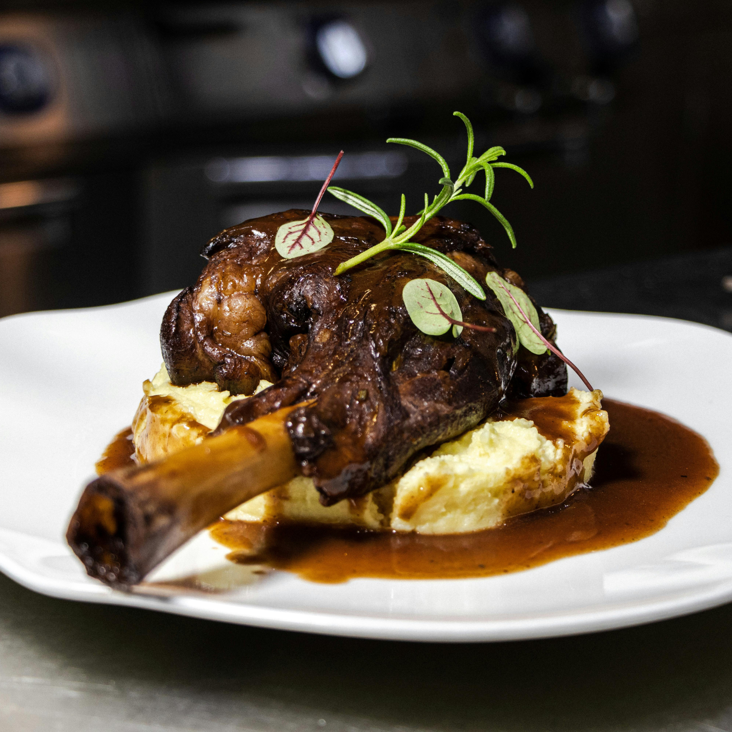 Braised lamb shank served on creamy mashed potatoes with rich brown gravy, garnished with fresh rosemary and microgreens on a white plate