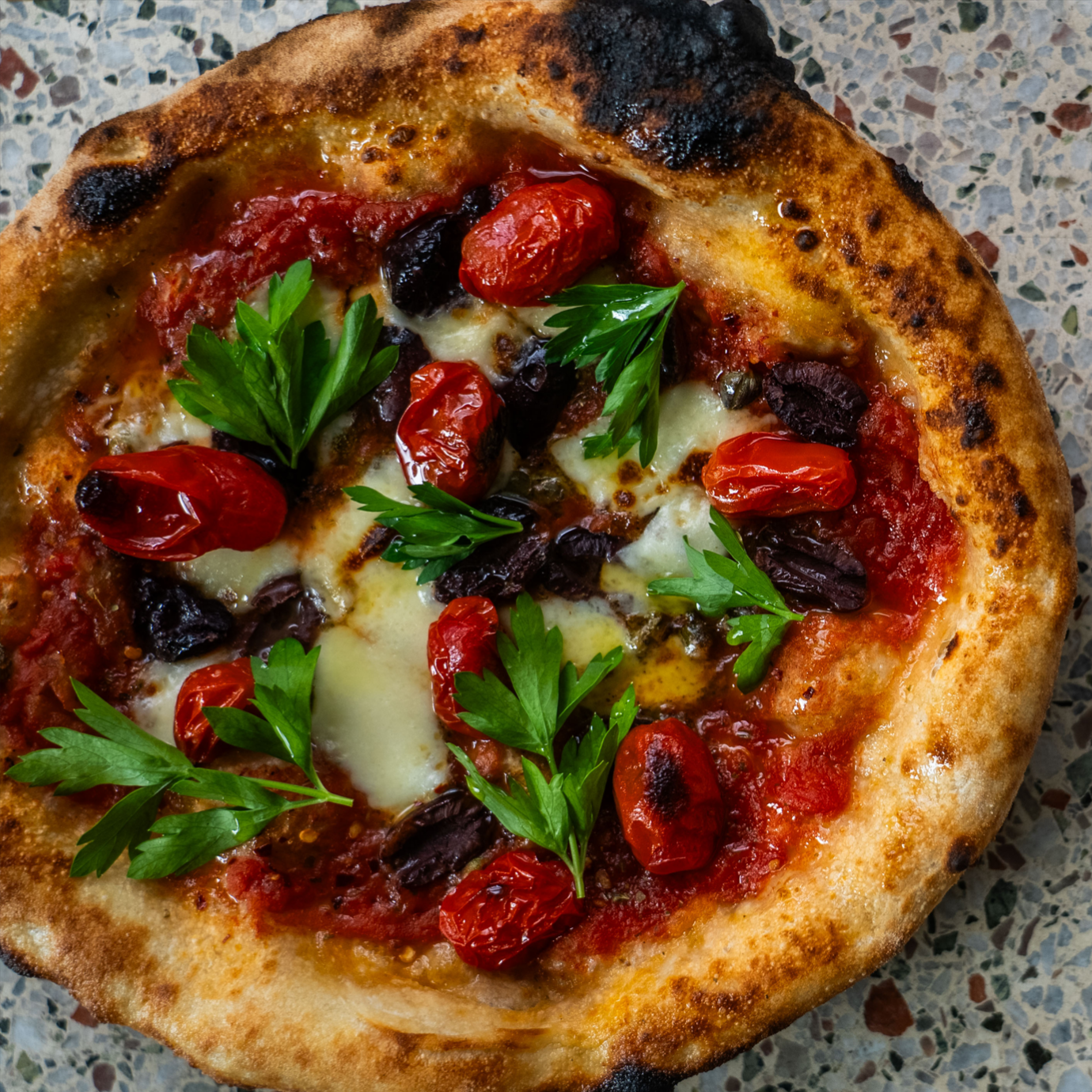 A freshly baked Neapolitan pizza with a puffy, charred crust, topped with roasted tomatoes and fresh parsley.
