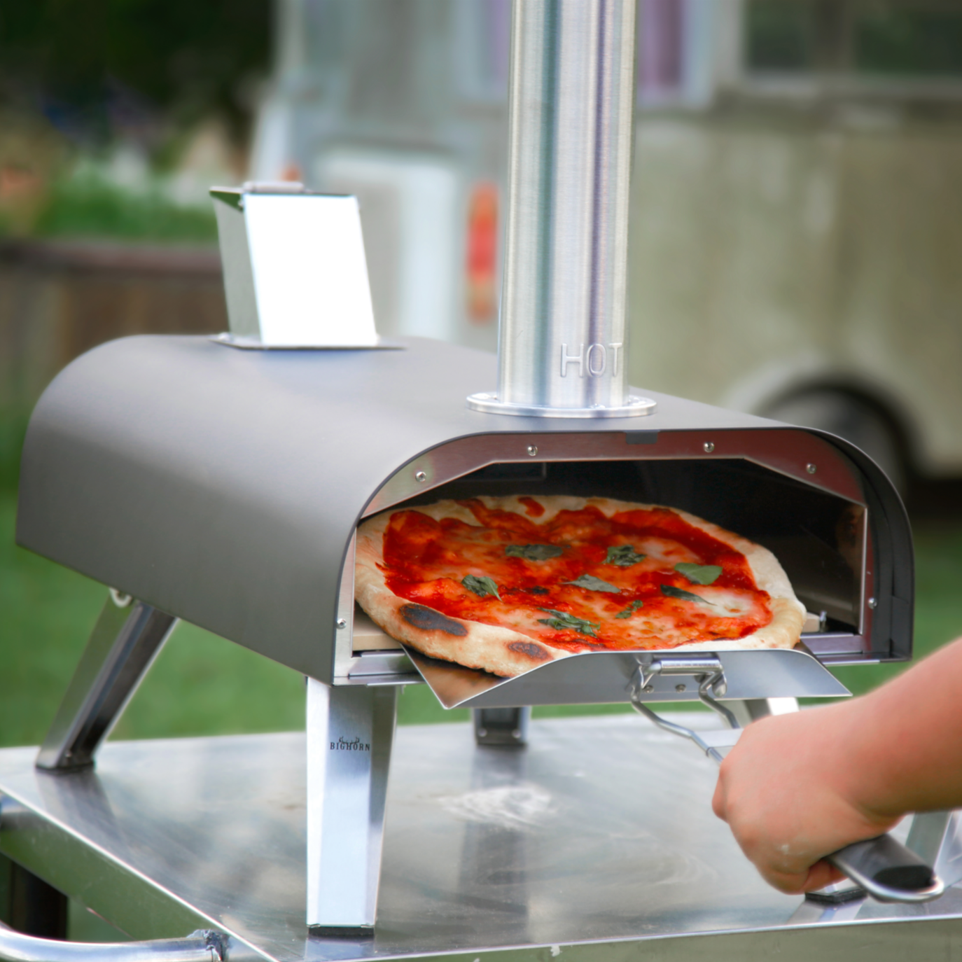 A pizza being placed into a stainless steel outdoor pizza oven.