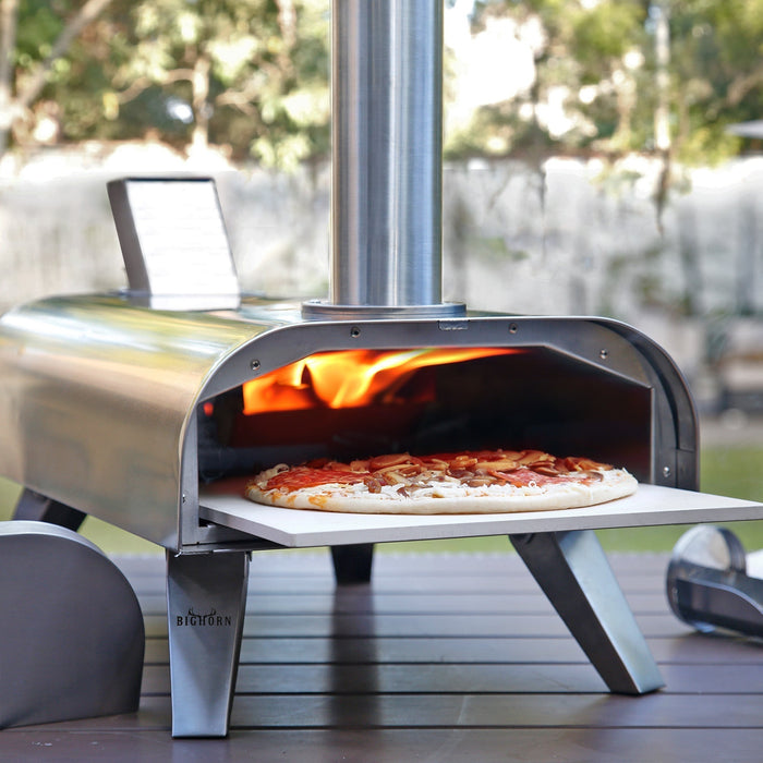 A stainless steel portable pizza oven with a pizza cooking on a stone inside, set on an outdoor wooden surface.