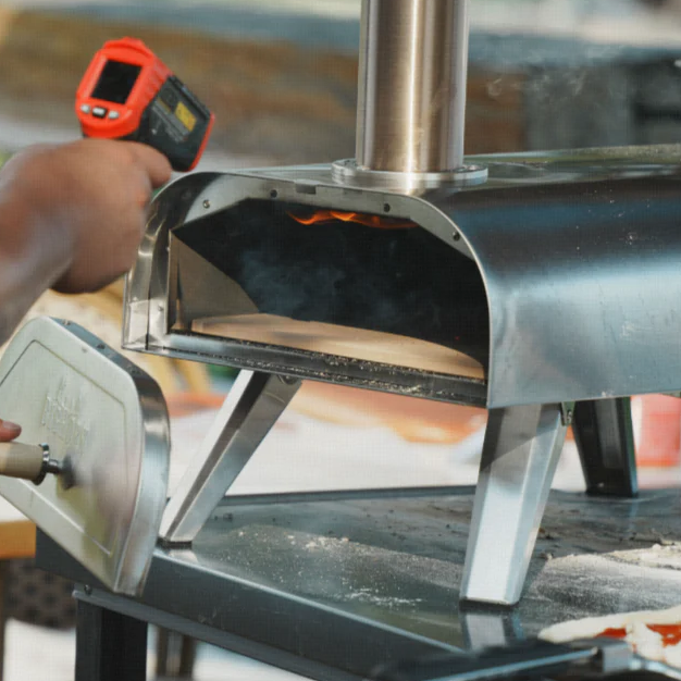 Person using a red infrared thermometer to check the heat inside a portable stainless steel pizza oven.