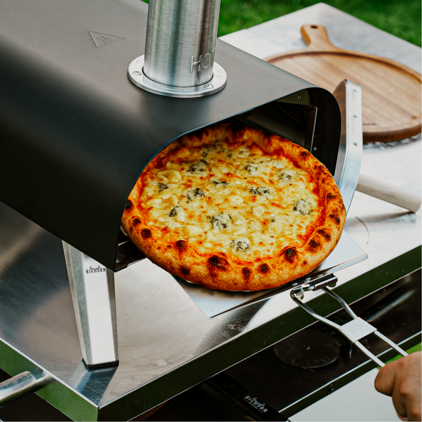 A cooked four-cheese pizza, featuring prominent blue cheese, emerges from a modern, black portable pizza oven outdoors.