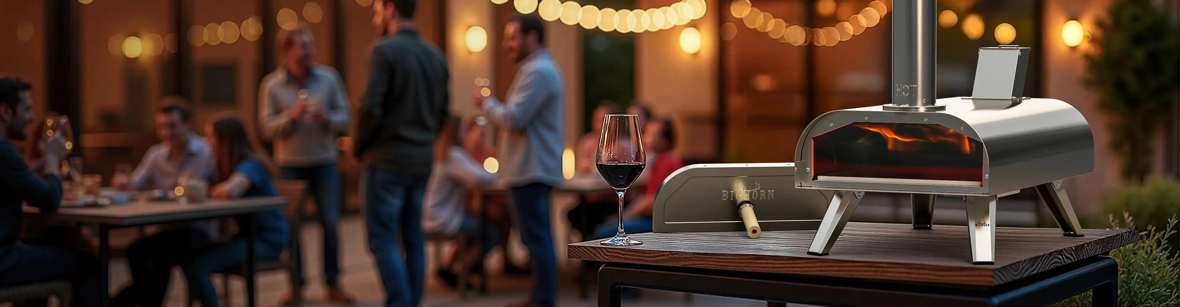 A bighorn outdoor stainless steel outdoor pizza oven on a wooden table at a social gathering, with a wine glass, people dining in the softly blurred background, and warm string lights overhead.