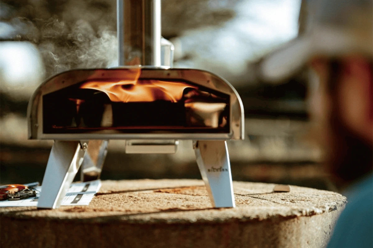 Portable outdoor bighorn pizza oven with fire inside on a wooden stump 
surface.